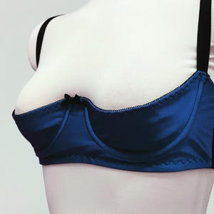 May include: A blue satin bra with black trim and straps. The bra has a classic design with a small bow detail at the center. The bra is displayed on a mannequin.