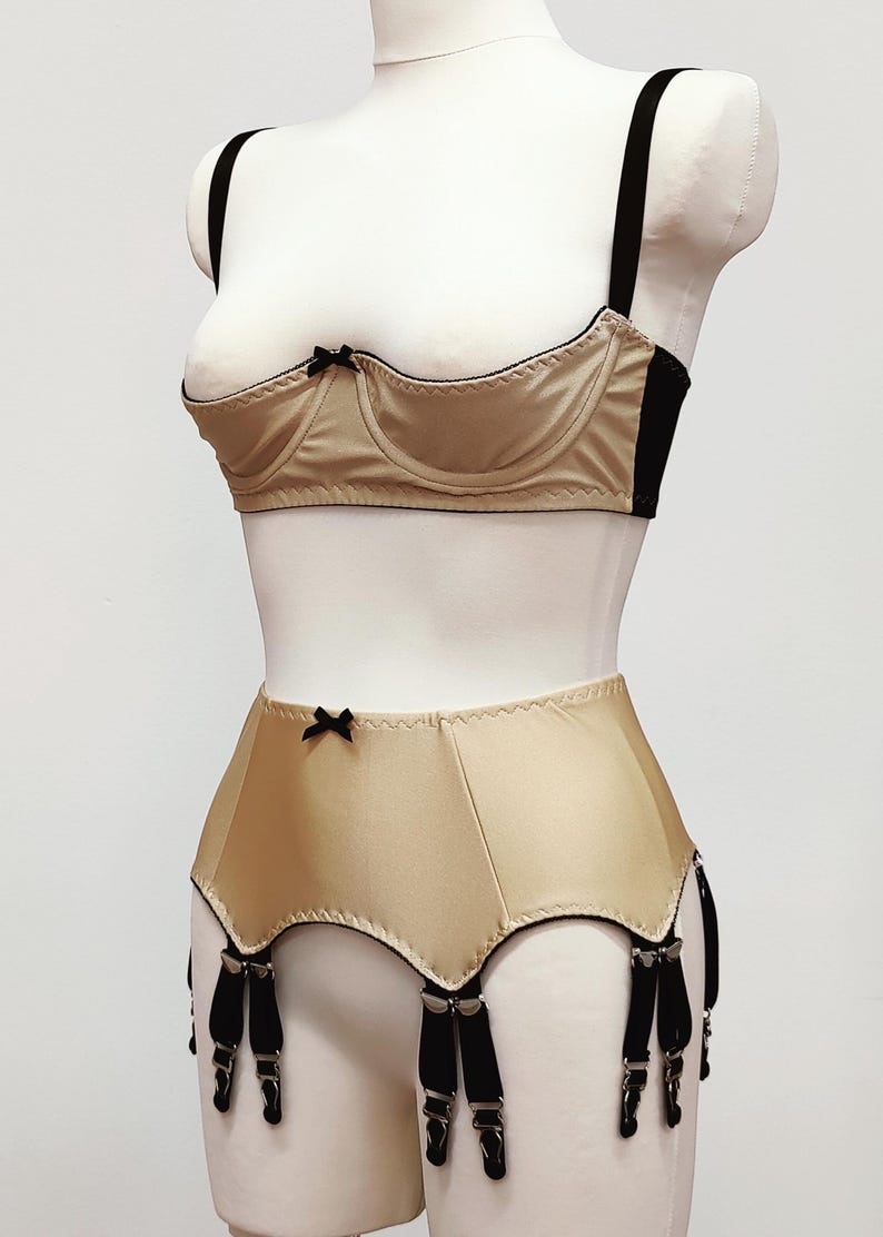 May include: A beige lingerie set on a mannequin. The set includes a bra with black trim and straps, and a high-waisted garter belt with black suspender straps. Small black bows adorn both pieces. The fabric has a satin sheen.