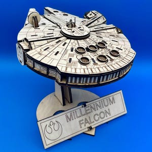 May include: A wooden Millennium Falcon model with intricate laser-cut details. The model sits on a stand, and a rectangular sign displays the name "Millennium Falcon" and a Rebel Alliance symbol. The model is made of light-colored wood.