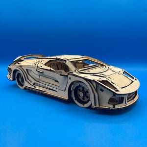 May include: A wooden model of a sports car, featuring intricate laser-cut details. The car is light brown, with visible wheel wells, a spoiler, and a detailed front grille. The model sits on a solid blue background.