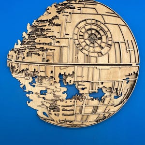 May include: A wooden Death Star wall art piece. The artwork is a detailed, laser-cut depiction of the iconic space station from the Star Wars franchise. The piece is made of light-colored wood and has intricate surface details.