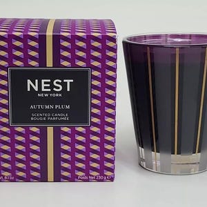 May include: A purple and gold striped glass candle jar with a matching box. The box has a geometric pattern and the text "NEST NEW YORK AUTUMN PLUM SCENTED CANDLE BOUGIE PARFUMEE" printed on it. The candle jar is 6.1 oz and the box is 230 g.