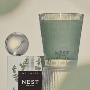 May include: A green glass candle with a white label that reads "NEST NEW YORK" and "WELLNESS". The candle is sitting on a white box with a green floral design.
