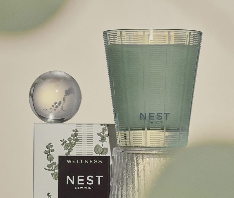 May include: A green glass candle with a white label that reads "NEST NEW YORK" and "WELLNESS". The candle is sitting on a white box with a green floral design.
