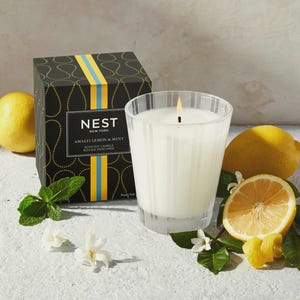May include: A black and gold candle jar with the text "NEST NEW YORK AMALFI LEMON & MINT SCENTED CANDLE BOUGIE PARFUMEE" on the front. The candle is lit and surrounded by lemons and greenery.