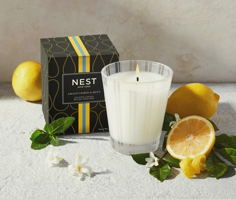 May include: A black and gold candle jar with the text "NEST NEW YORK AMALFI LEMON & MINT SCENTED CANDLE BOUGIE PARFUMEE" on the front. The candle is lit and surrounded by lemons and greenery.