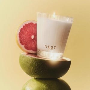 May include: A white candle in a clear glass jar with a gold label that reads "NEST NEW YORK". The candle is sitting on top of a halved grapefruit and a green pomelo.