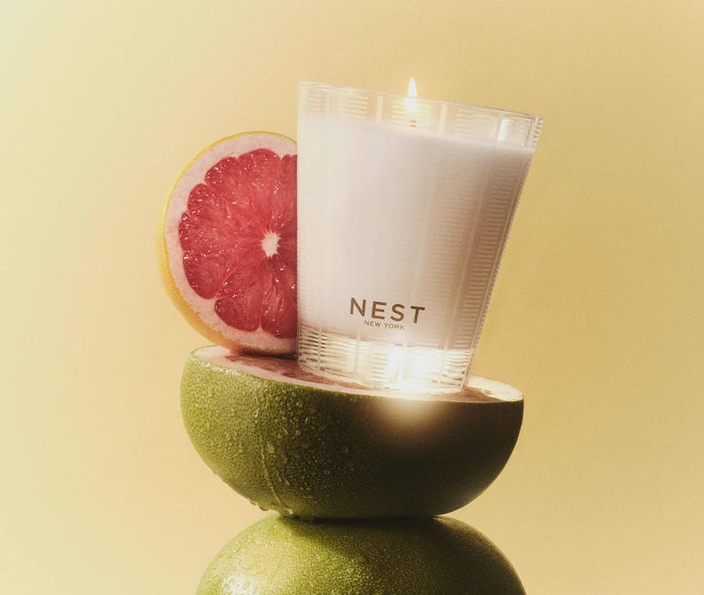 May include: A white candle in a clear glass jar with a gold label that reads "NEST NEW YORK". The candle is sitting on top of a halved grapefruit and a green pomelo.