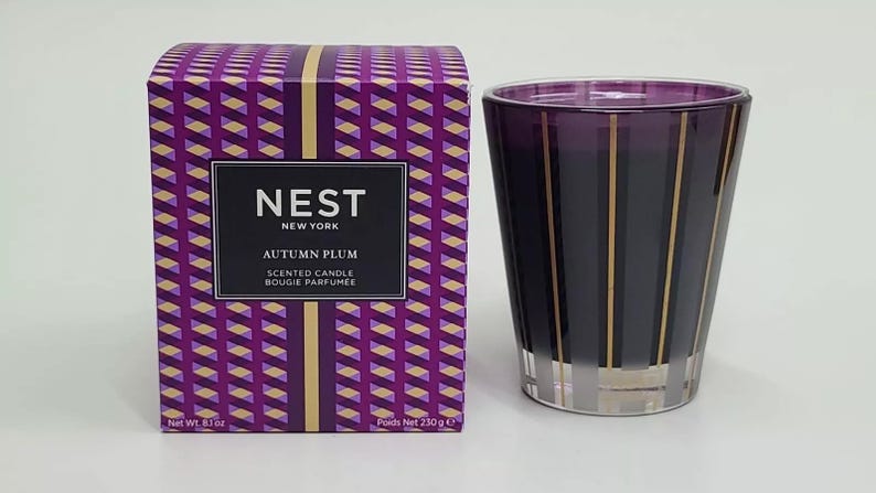 May include: A purple and gold striped glass candle jar with a matching box. The box has a geometric pattern and the text "NEST NEW YORK AUTUMN PLUM SCENTED CANDLE BOUGIE PARFUMEE" printed on it. The candle jar is 6.1 oz and the box is 230 g.