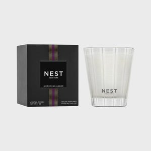 May include: A black and gold candle box with the text "NEST NEW YORK" and "MOROCCAN AMBER". The candle is in a clear glass jar with a white wax and the text "NEST NEW YORK" on the front.