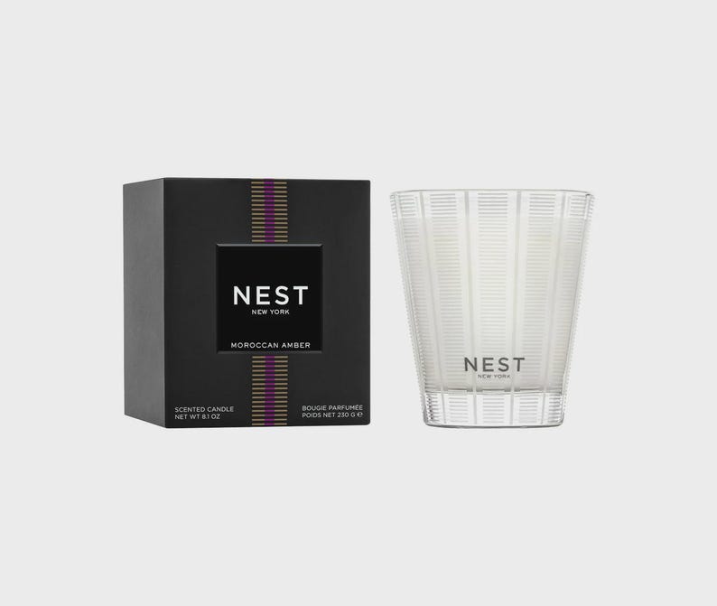 May include: A black and gold candle box with the text "NEST NEW YORK" and "MOROCCAN AMBER". The candle is in a clear glass jar with a white wax and the text "NEST NEW YORK" on the front.