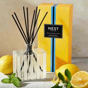 May include: A clear glass bottle with black reeds and a yellow and blue label that reads "NEST NEW YORK AMALFI LEMON & MINT REED DIFFUSER DIFFUSEUR DE PARFUM". The bottle is sitting on a white surface with lemons and greenery.