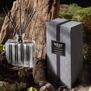May include: A gray and white striped glass diffuser bottle with black reeds and a gray box with the text "NEST NEW YORK WILDERNESS CHARCOAL WOOD REED DIFFUSER DIFFUSEUR DE PARFUM" on the front.
