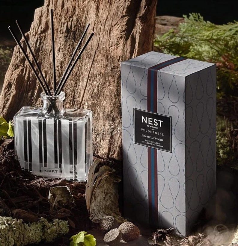 May include: A gray and white striped glass diffuser bottle with black reeds and a gray box with the text "NEST NEW YORK WILDERNESS CHARCOAL WOOD REED DIFFUSER DIFFUSEUR DE PARFUM" on the front.