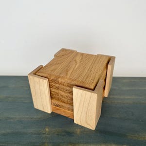 May include: A wooden coaster set with a natural wood grain finish. The set includes a stack of square coasters held within a light-coloured wooden holder. The holder has four corner pieces that secure the coasters. The coasters and holder are made of different types of wood.