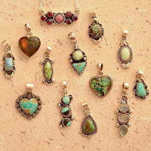 Puede incluir: Una colección de colgantes de plata con diversos diseños de piedras preciosas. Los colgantes presentan formas de corazón, ovaladas y de lágrima, con piedras turquesas, verdes y rosas. Algunos tienen detalles de plata.