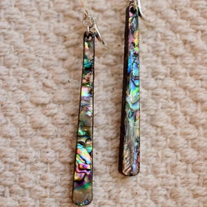 May include: Two long, rectangular earrings featuring iridescent abalone shell inlays. The earrings have a dark brown border and silver ear wires. The abalone shell displays a range of colours, including green, blue, and purple.