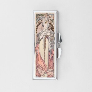 May include: A rectangular metal pill case featuring a decorative Art Nouveau illustration of a figure in a flowing cream and pink gown. The case has a polished silver-tone frame with a side hinge and a latch mechanism on the right edge.