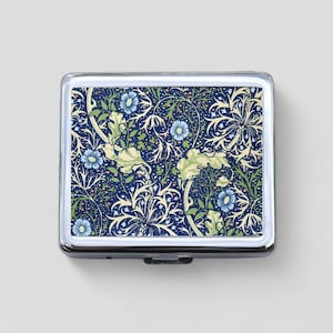 May include: A silver metal pill box with a floral design. The design features blue, green, and white flowers on a dark blue background.