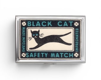 Puede incluir: Una caja de cerillas rectangular de metal con una etiqueta de estilo vintage que muestra una ilustración de un gato negro sobre fondo crema. El texto dice BLACK CAT, SAFETY MATCH, MADE IN POLAND e IMPREGNATED, con un borde negro y detalles azules.