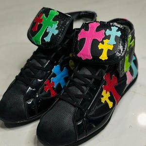 Puede incluir: Un par de zapatillas altas negras con diseños de cruces coloridas. Las zapatillas tienen un acabado brillante y cierre de cordones. Los diseños de cruces son de varios colores, incluyendo verde, rosa, azul, amarillo y rojo.