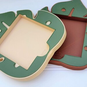 May include: Two decorative frames with a leaf-like design. One frame is beige with green trim, and the other is brown with green trim. Both frames have a rectangular interior and rounded edges. The frames are likely made of plastic.