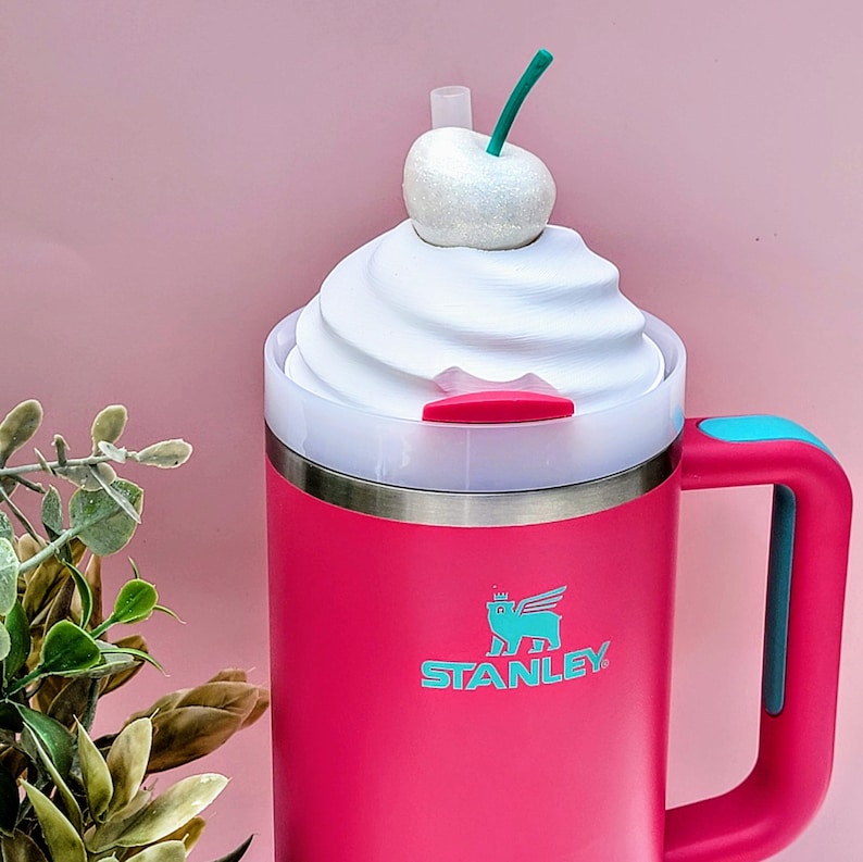 May include: A pink Stanley travel mug with a white whipped cream-like lid, topped with a glittery white cherry and green stem. The mug has a teal handle and the Stanley logo in teal. A clear straw is inserted into the lid.