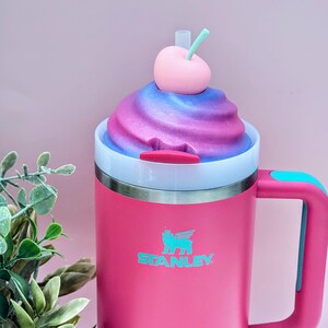 May include: A pink Stanley mug with a white and silver rim, a blue handle, and a decorative top resembling a swirl of pink and blue frosting, topped with a pink cherry and a green stem. The Stanley logo is visible on the mug.