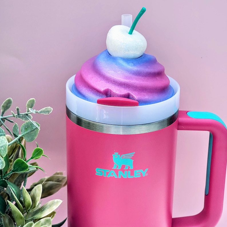 May include: A pink Stanley tumbler with a handle and a white lid. The lid is topped with a colorful, whipped cream-like design and a white cherry with a green stem. The Stanley logo is displayed in teal on the front.