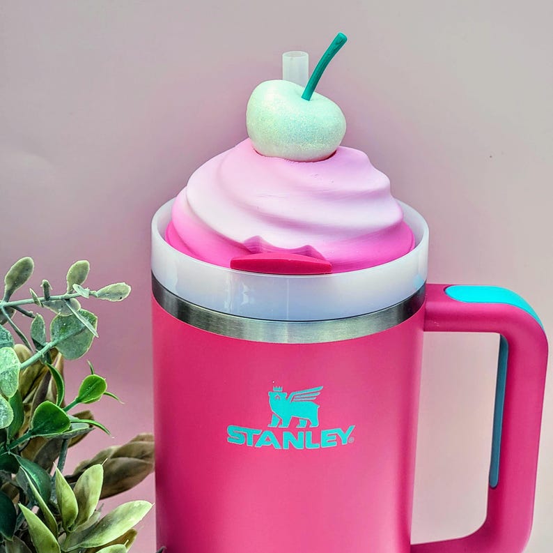 May include: A pink Stanley mug with a handle and a white and silver rim. The mug is topped with a swirl of pink and white frosting and a green cherry. The Stanley logo is printed in teal on the front.