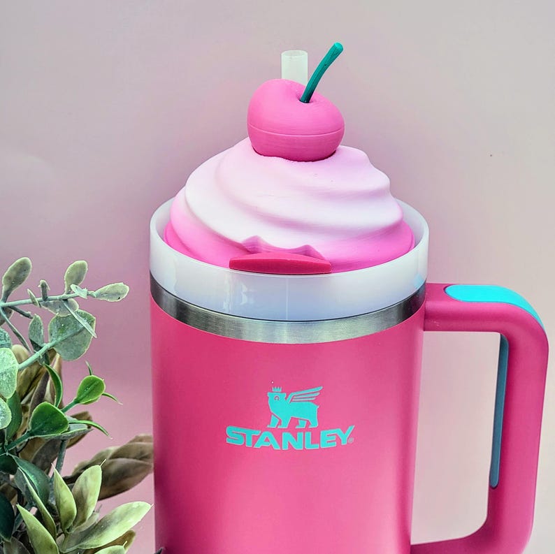 May include: A pink Stanley mug with a white and pink swirl design on top, resembling whipped cream. A pink cherry with a green stem sits on top. The mug has a handle with a blue accent and the Stanley logo in teal.
