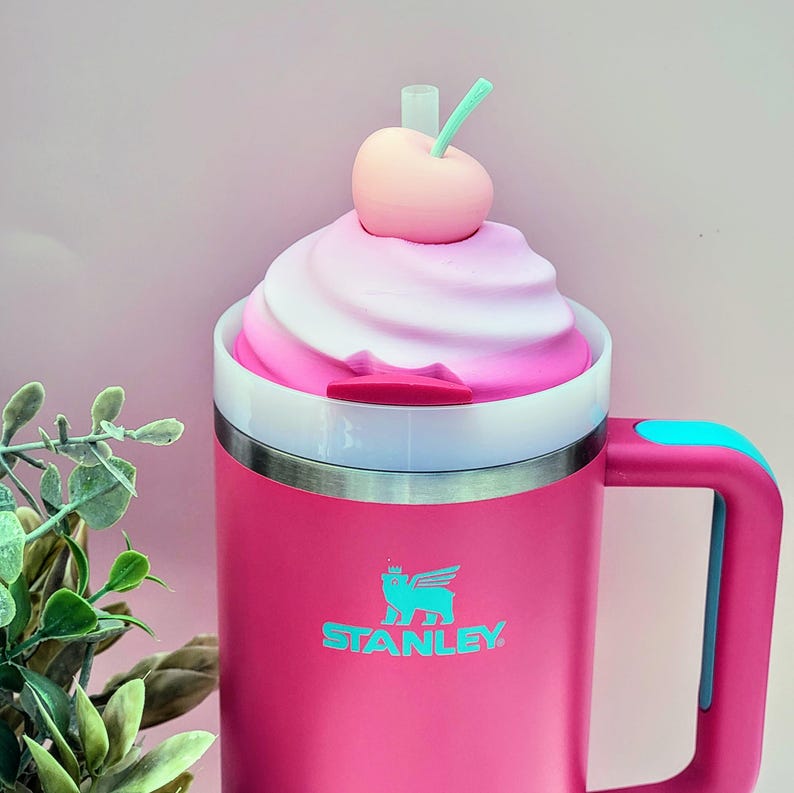 May include: A pink Stanley mug with a white and pink swirl design on top, topped with a pink cherry and a green straw. The mug has a handle with a blue accent and the Stanley logo in teal.