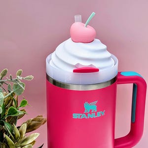 May include: A pink Stanley travel mug with a white whipped cream-like lid, topped with a pink cherry and green stem. The mug has a teal handle and the Stanley logo. The mug is designed to look like a milkshake.