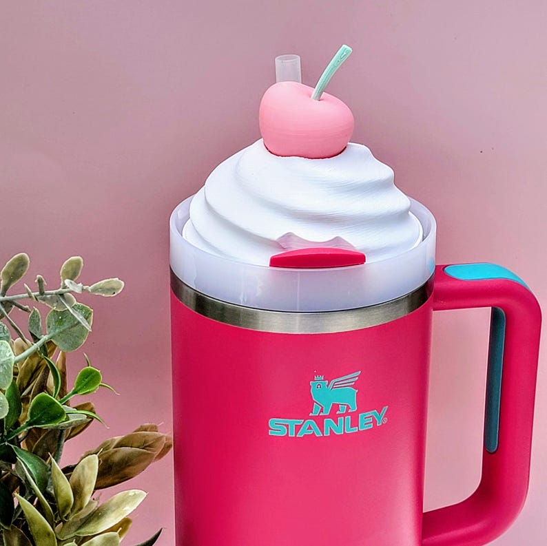 May include: A pink Stanley travel mug with a white whipped cream-like lid, topped with a pink cherry and green stem. The mug has a teal handle and the Stanley logo. The mug is designed to look like a milkshake.