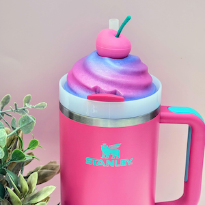 May include: A pink Stanley mug with a white rim and a blue handle. The mug is topped with a swirl of pink and purple foam, and a pink cherry with a green stem. The Stanley logo is in teal.
