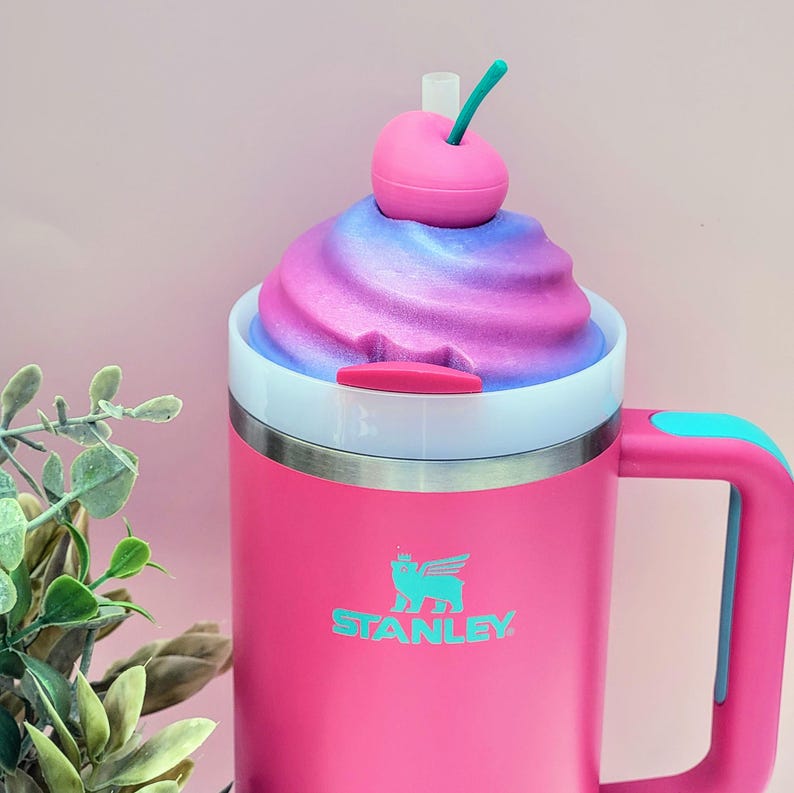 May include: A pink Stanley mug with a white rim and a blue handle. The mug is topped with a swirl of pink and purple foam, and a pink cherry with a green stem. The Stanley logo is in teal.
