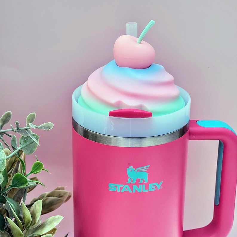 May include: A pink Stanley insulated mug with a handle and a decorative top resembling a swirl of pastel-colored soft serve ice cream, topped with a pink cherry and a green stem. The mug has a silver rim and the Stanley logo.