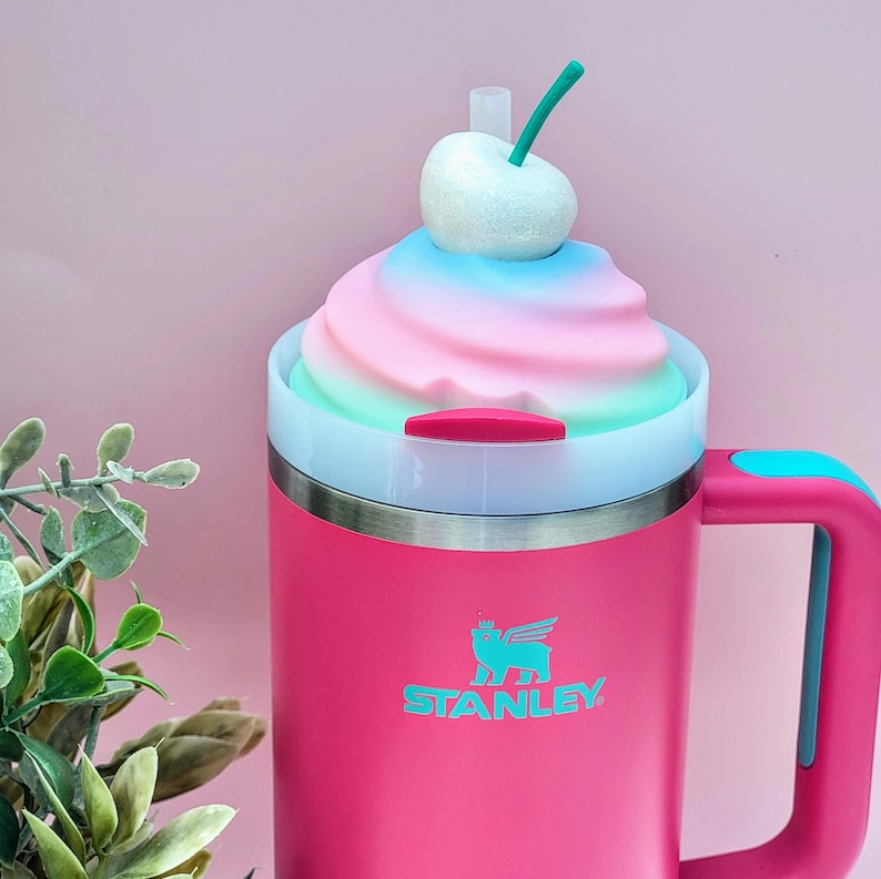 May include: A pink Stanley tumbler with a handle and a decorative top resembling a swirl of pastel-colored ice cream, topped with a white cherry and green stem. The tumbler has a teal handle and the Stanley logo.