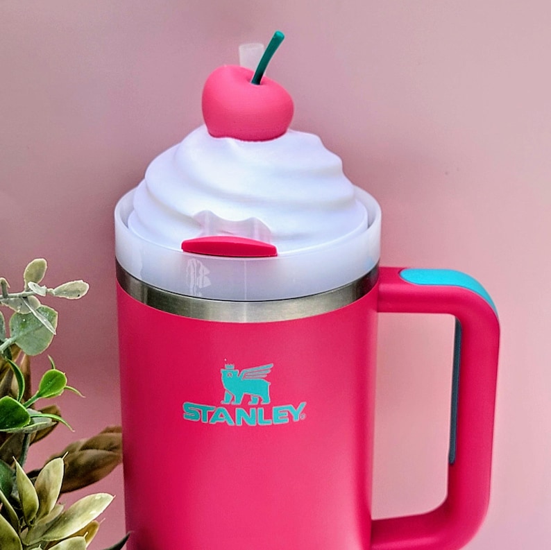 May include: A pink Stanley travel mug with a white whipped cream-like lid, topped with a pink cherry and green stem. The mug has a teal handle accent and the Stanley logo in teal. The mug is designed to look like a milkshake.