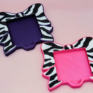 May include: Two decorative bow-shaped trays. One tray is purple, the other is pink. Both have a zebra print design in black and white on the bow. The trays are on a pink background.
