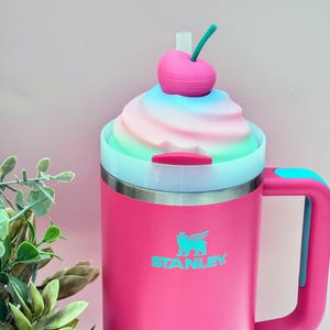 May include: A pink Stanley travel mug with a whimsical design. The mug features a decorative swirl of pastel colors and a cherry on top. The handle and accents are teal. The Stanley logo is visible on the front.