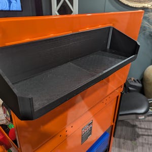 May include: A black rectangular storage tray with an open top and angled side walls, mounted onto the side of a bright orange metal tool chest. The tray features a textured surface and a wide, flat base designed for holding various small items.