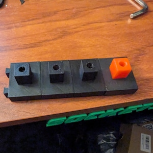 May include: A black 3D-printed component with four square sections. Three sections have a circular hole, and the fourth is an orange cube. The component is on a wooden surface.
