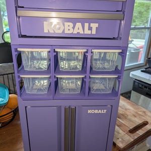 May include: A purple Kobalt storage unit featuring a top drawer with silver handles, a middle section with six clear plastic bins, and a bottom cabinet with two doors. The brand name Kobalt is printed in white on the drawer and the cabinet door.