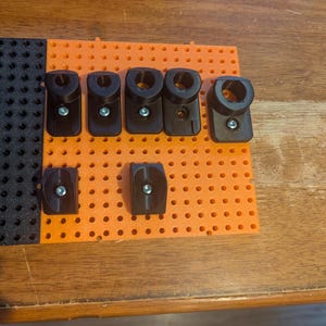 May include: A bright orange pegboard panel featuring a grid of circular holes, with seven black plastic mounting brackets attached to the surface using silver metal screws. The brackets vary in shape, with some featuring circular openings.