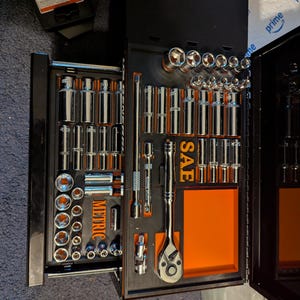 May include: A black toolbox with an open drawer revealing a comprehensive set of chrome sockets and wrenches. The drawer is labeled "METRIC" and "SAE" with orange accents. The tools are neatly organized.