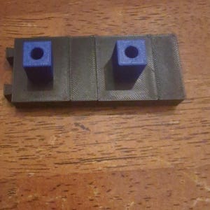 May include: A 3D-printed, dark gray object with two blue square blocks. Each block has a circular hole in the center. The object is on a wooden surface.