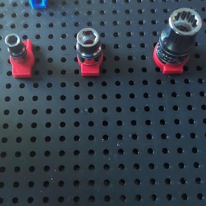 May include: A collection of socket wrench attachments in various sizes. The sockets are silver and black, mounted on red bases. A blue plastic piece is also present. The items are arranged on a black pegboard.