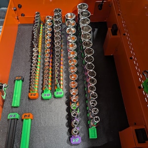 May include: A collection of chrome-plated metal socket sets organized on vertical rails inside an orange tool chest. The rails are labeled 1/4, 3/8, and 1/2, featuring green, orange, and purple plastic bases. The sockets vary in size and depth.