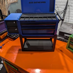May include: A blue U.S. General tool chest with a hinged lid, multiple drawers, and a side storage compartment. The unit features black handles and trim, with the brand name U.S. General printed on the lid interior and the front of the main cabinet.